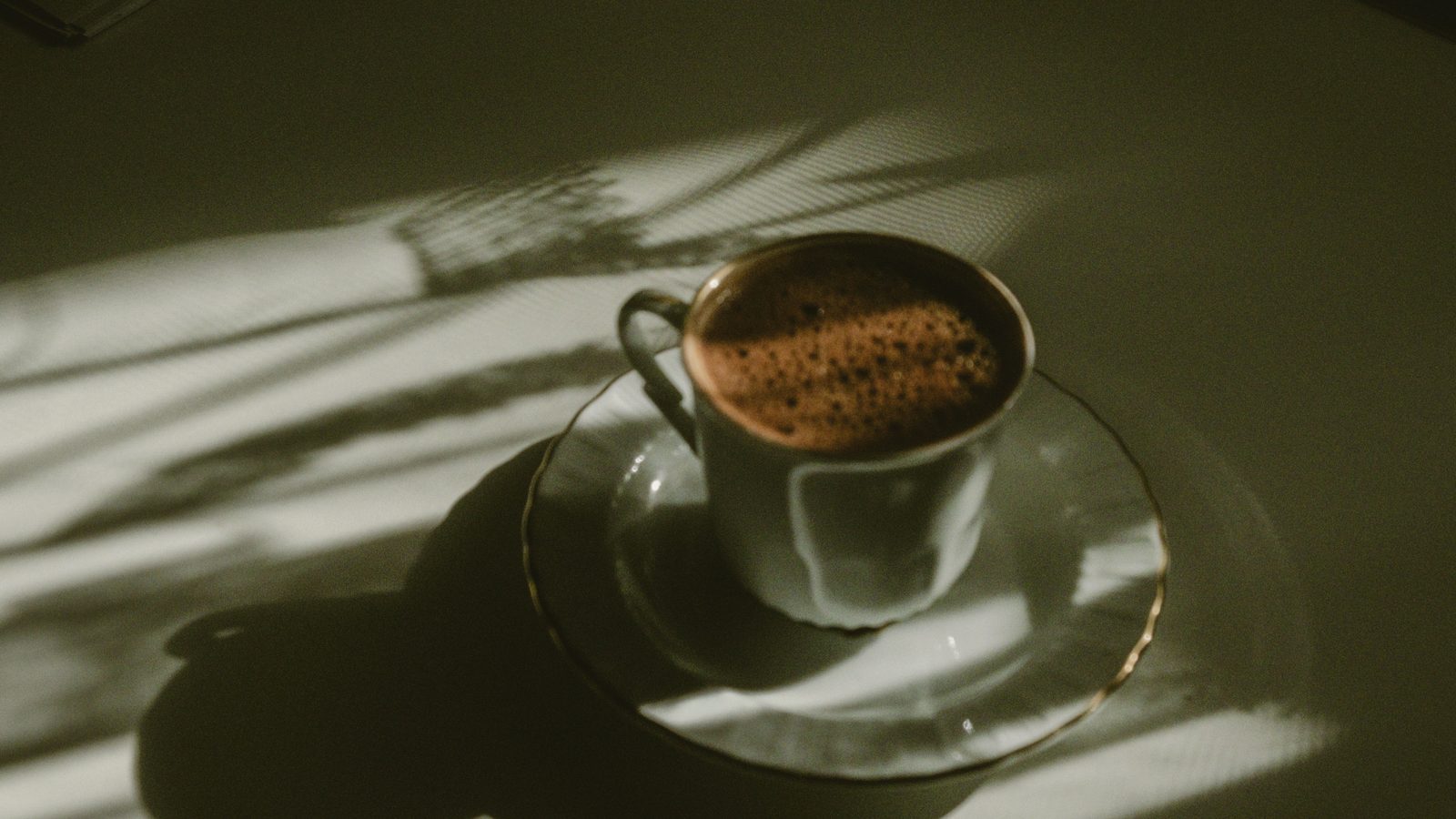 A cup of coffee on a pale table, lit by dappled early light through a window. Most of the frame is in shadow.