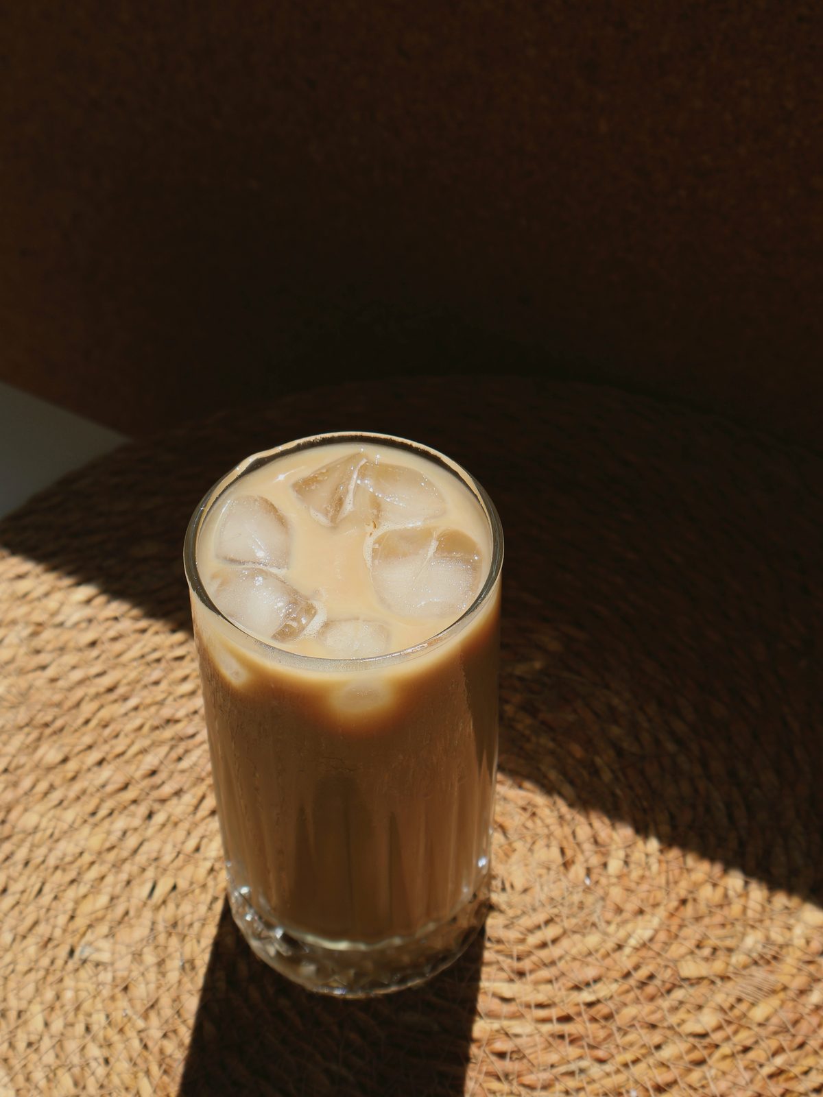 Iced coffee with milk in a tall glass on a woven mat, in hard side light.
