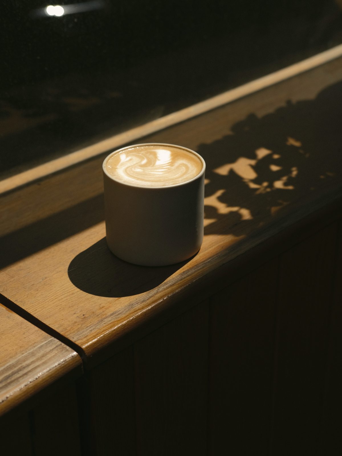 A latte in a matte ceramic tumbler on a wooden sill, dappled sunlight behind it.