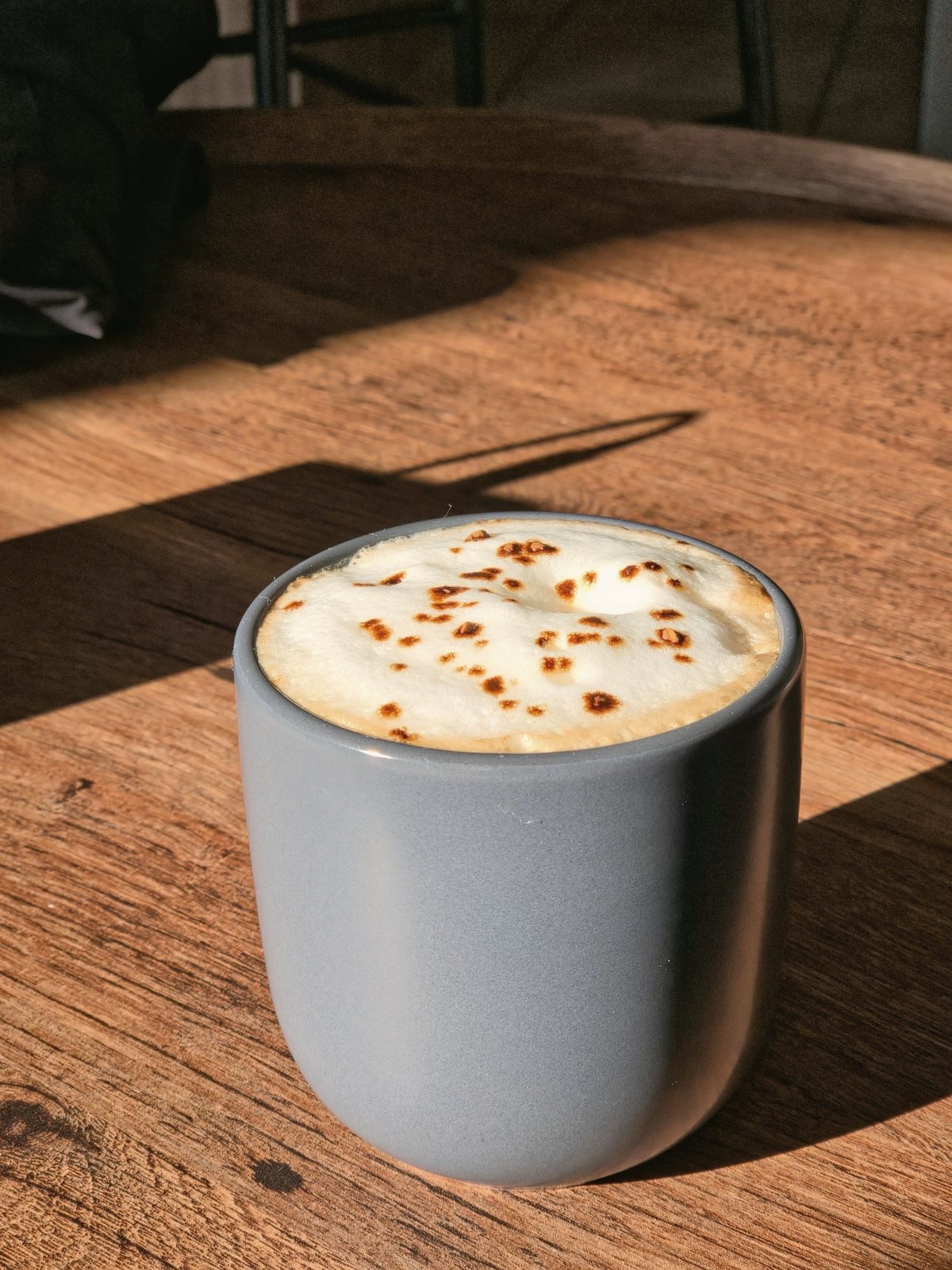 A grey stoneware cup of foamed coffee on an oak table in low sun.