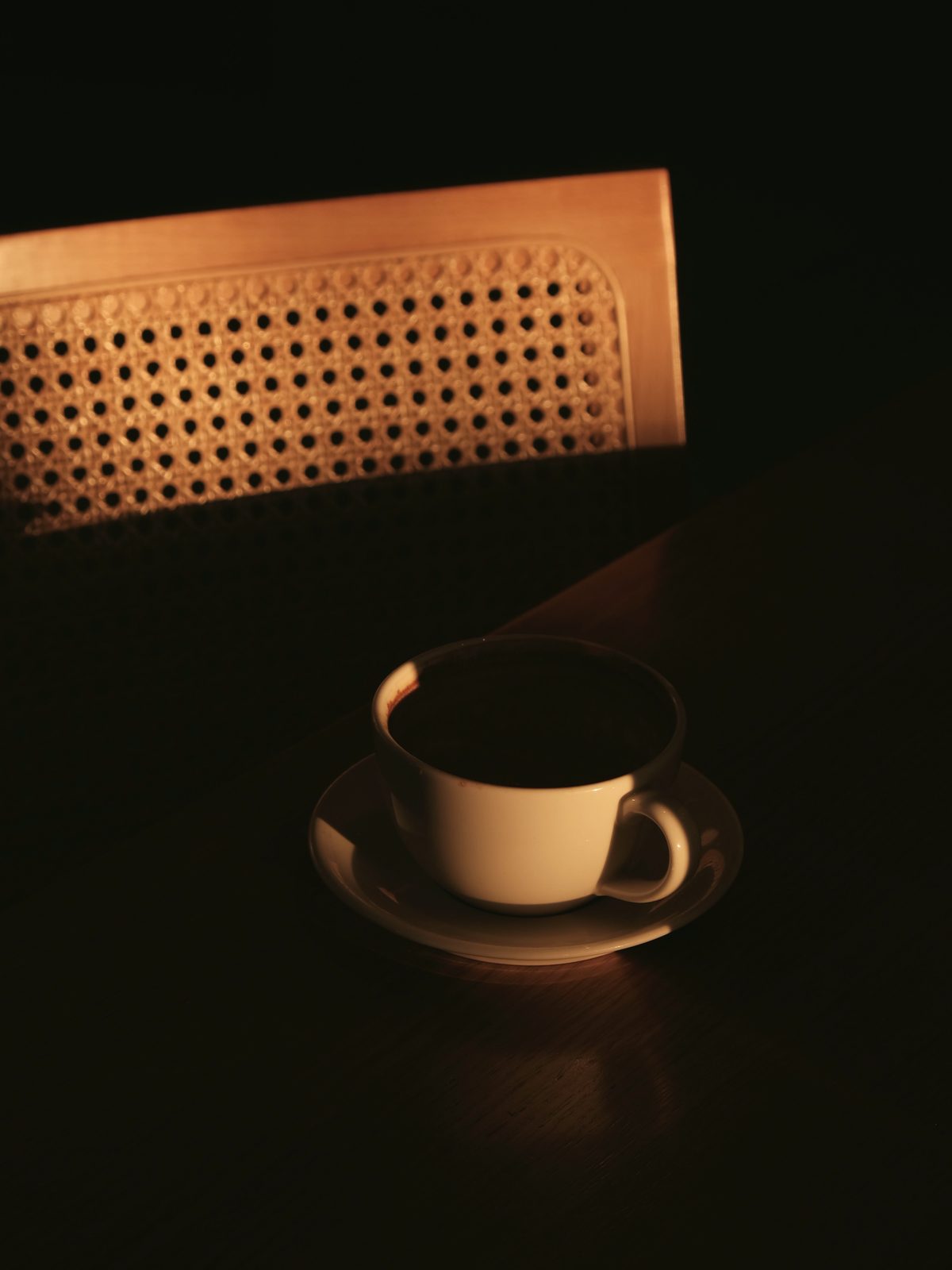 A cup of dark tea on a saucer in near darkness, a cane chair catching the last light.