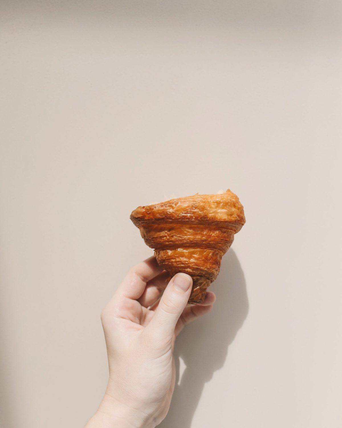 A hand holding a croissant up against a plaster wall in morning sun.