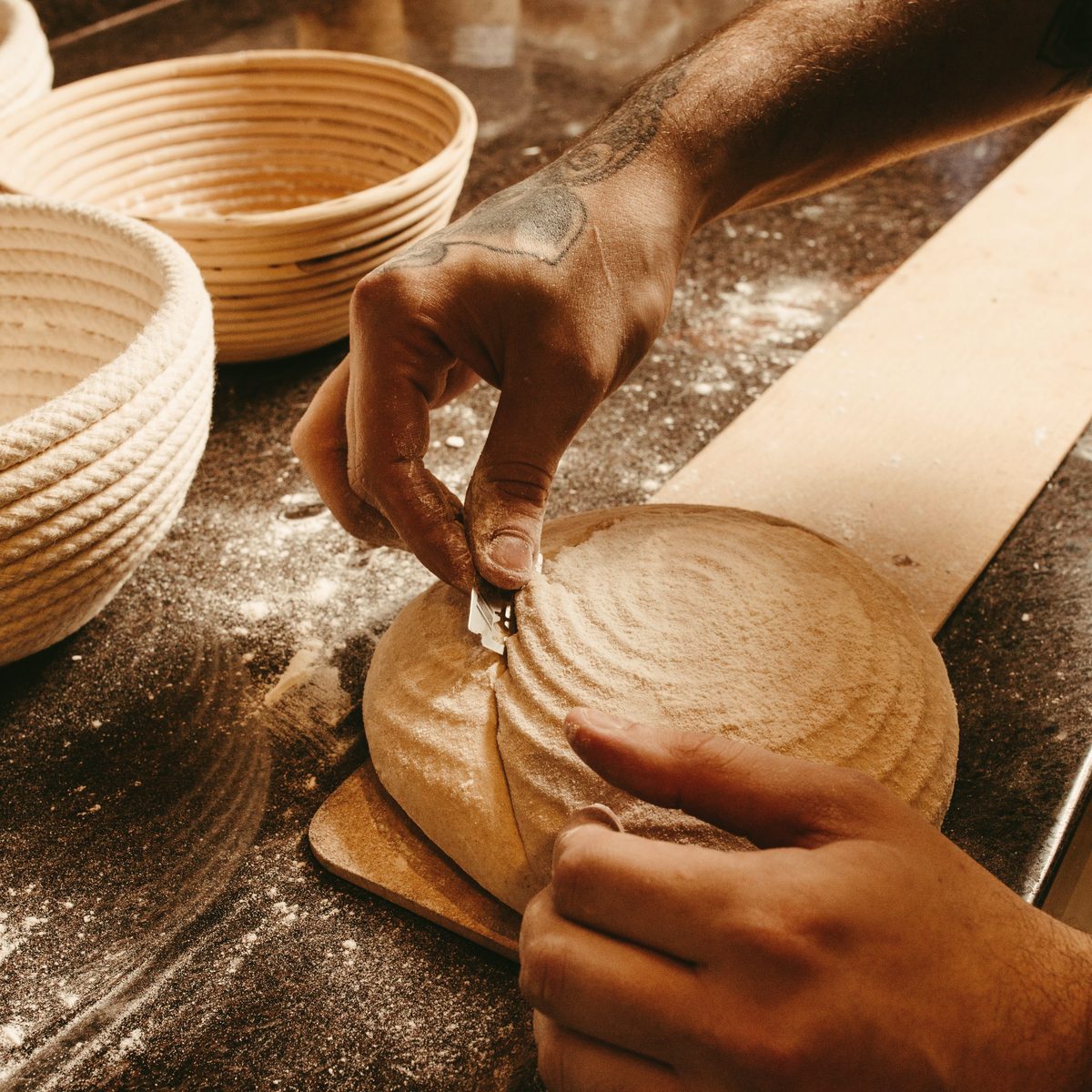 A baker's hands scoring a round loaf beside bannetons, under warm working light.