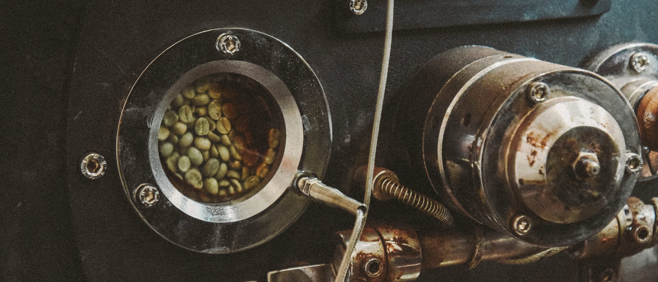 Green coffee visible through the sight glass of a steel roasting drum.