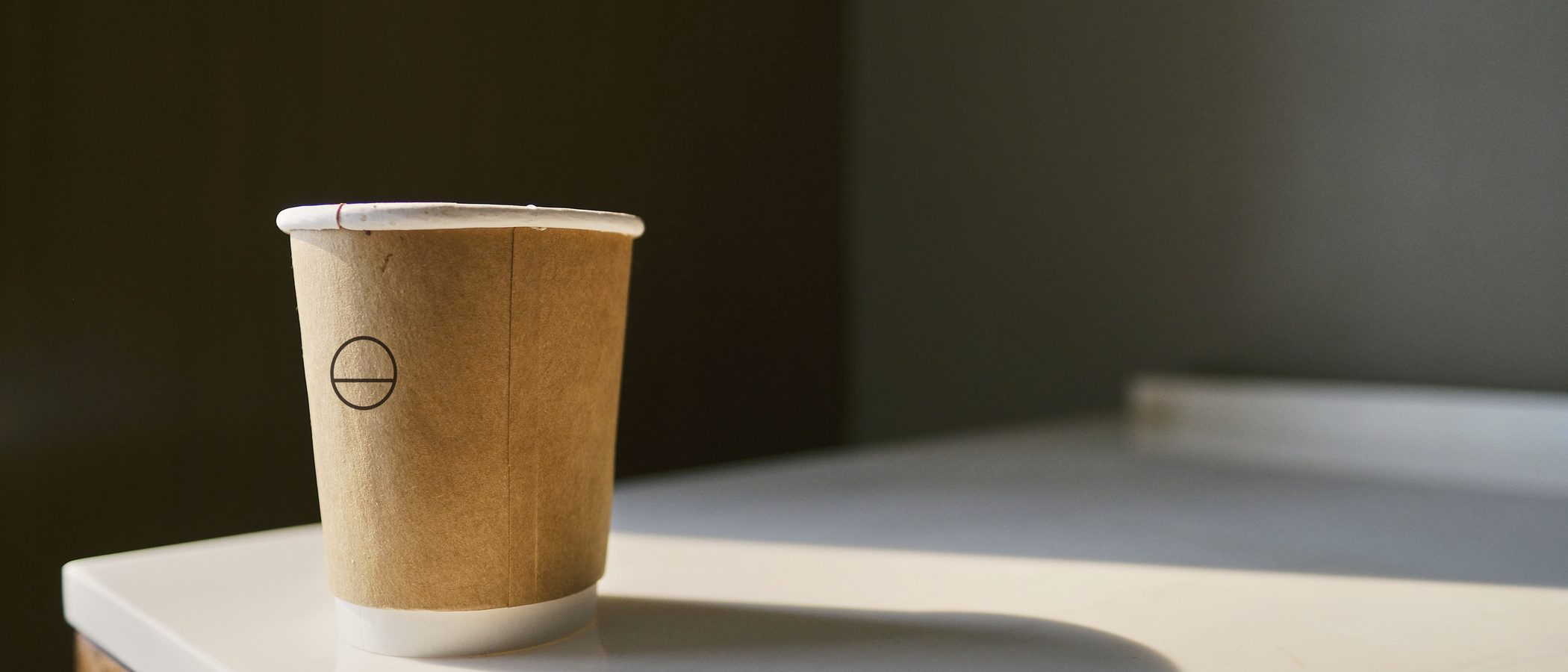 A kraft takeaway cup printed with the Morrow mark, on a white counter in hard morning light, the wall behind it in shadow.